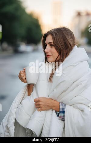 Seitenbokeh-Porträt einer Frau, die in eine Decke gehüllt ist und Kaffee über dem Hintergrund der Stadt trinkt. In den frühen Morgenstunden. Stockfoto