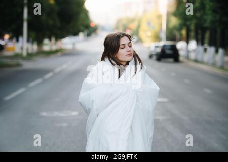 Eine glückselige, gemütliche Frau, die am frühen Morgen in eine Decke auf einer leeren, nassen Stadtstraße gehüllt wurde. Zwischen Baumreihen. Weiche Decke. Stockfoto