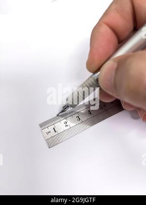 Hand mit einem mechanischen Metallstift, der eine gerade Linie mit einem metallischen Millimeterlineal auf einem leeren Blatt Papier macht, selektiver Fokus im Lineal, V Stockfoto