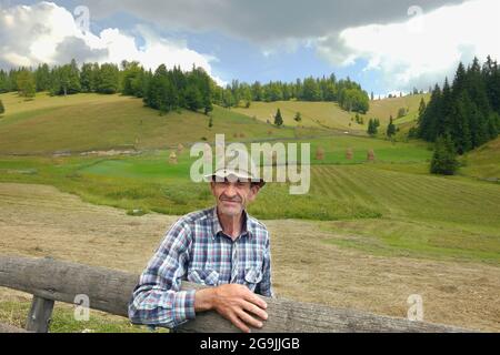 GARDA DE SUS, RUMÄNIEN - 04. AUGUST 2015: Der Seniorbauer schaut mich an und lehnt sich mit der Hand an den Holzzaun der Landschaft im Apuseni-Gebirge Stockfoto