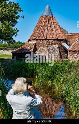 Ältere Frau, die am sonnigen Sommertag, East Lothian, Schottland, Großbritannien, Fotos von der historischen, skurrilen alten Preston Mill macht, die sich im Mühlenteich widerspiegelt Stockfoto