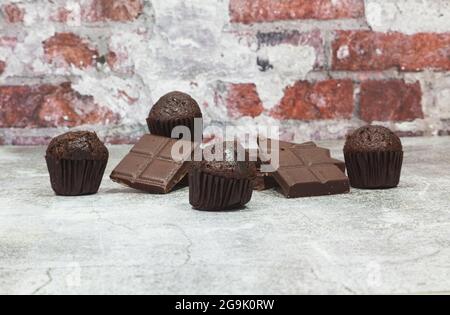 Auf einer grauen Oberfläche vor einer Ziegelwand sind mehrere Stücke einer dunklen Schokoladenriegel aufgestapelt und einige Schokoladen-Cupcakes. Stockfoto