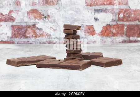 Auf einer grauen Fläche vor einer Ziegelwand befindet sich ein Turm aus Unzen dunkler Schokolade. Um ihn herum befinden sich mehr Stücke einer Schokoladenriegel. Stockfoto