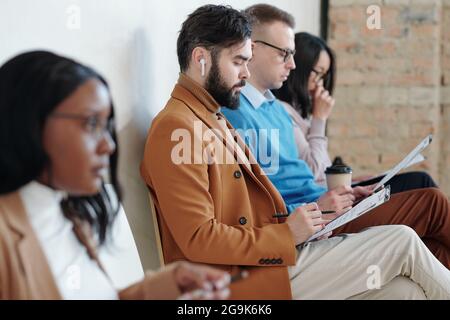 Eine Gruppe ernsthafter junger interkultureller Menschen, die in Reihe gegen die Wand sitzen Stockfoto