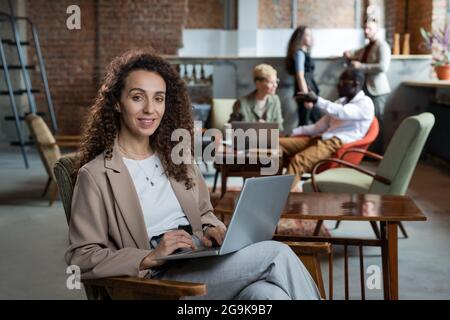 Glückliche junge Geschäftsfrau mit Laptop sitzt im Sessel in der Arbeitsumgebung Stockfoto