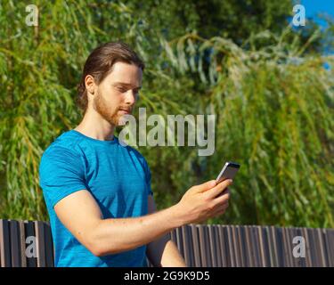 Junger, hübscher Sportler, der die Fitness-App auf einem modernen Smartphone nutzt, während er im Park trainiert Stockfoto