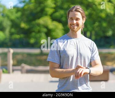 Positiver junger, gutaussehender Sportler in Kopfhörern mit Fitnessarmband beim Training im Freien Stockfoto