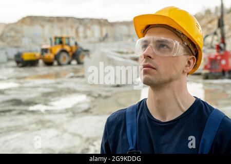 Junger seriöser Geschäftsmann in Hardhut und Brille, der am Bauplatz steht Stockfoto