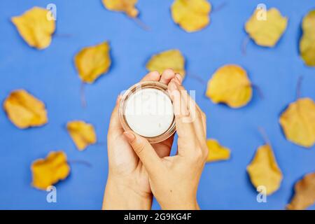 Herbstcreme über Herbstblättern. Kurzer Schuss weiblicher Hände, die eine feuchtigkeitsspendende Gesichtscreme auf einem hellen, farbenfrohen Herbsthintergrund mit Orange halten Stockfoto