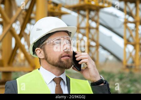 Junger Ingenieur in Arbeitshelm und Schutzbrille im Freien stehend und im Gespräch auf dem Handy Stockfoto