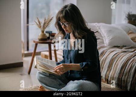 Junge, fokussierte asiatische Frau, die eine Brille trägt und ein Buch liest, während sie in Lotuspose auf dem Boden im gemütlichen Wohnzimmer sitzt Stockfoto