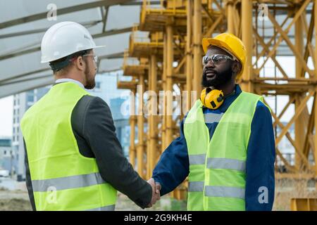 Zwei Ingenieure in Arbeitshelmen und reflektierender Kleidung schütteln sich die Hände und grüßen sich gegenseitig, während sie auf der Baustelle stehen Stockfoto