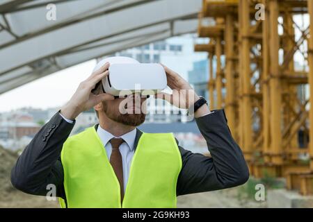 Ein reifer Ingenieur blickt durch die Virtual-Reality-Brille auf das zukünftige Projekt, während er auf der Baustelle steht Stockfoto