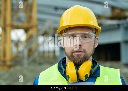 Nahaufnahme eines Bauarbeiters im Arbeitshelm und in Kopfhörern, der die Kamera im Freien betrachtet Stockfoto