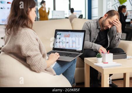 Rückansicht Video von jungen Geschäftsfrau sitzen auf Couch im Büro mit Laptop-Computer mit Grafiken, während Kollegen arbeiten im Hintergrund. Multiethnische Mitarbeiter planen ein neues Finanzprojekt Stockfoto