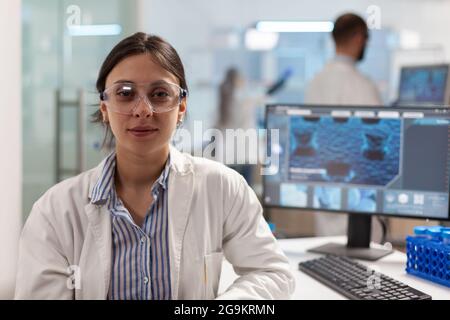 Wissenschaftler mit Laborkittel sitzt im Labor und schaut lächelnd auf die Kamera, während ein Kollege im Hintergrund am Computer arbeitet. Chemiker untersucht die Evolution von Viren mithilfe von Hightech für die wissenschaftliche Forschung, Impfstoff. Stockfoto