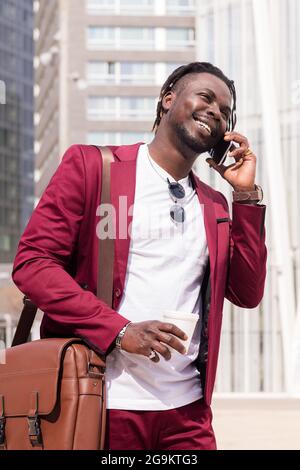 Vertikales Porträt eines lächelnden afrikanischen Geschäftsmannes, der durch die finanzielle Innenstadt der Stadt läuft und am Telefon mit einem Kaffee in der Hand telefoniert, Konzept Stockfoto