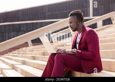 Der elegante schwarze Geschäftsmann arbeitet mit seinem Laptop, der auf einer Treppe in der Stadt sitzt, mit Technologie und Remote-Work-Konzept Stockfoto