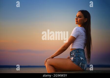 Junge Frau, die im Profil auf einem Pier sitzt und den Sonnenuntergang beobachtet Stockfoto