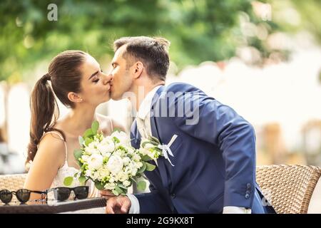 Fröhliches Paar sitzt und küsst sich nach der Hochzeit. Die Braut hält einen eleganten Strauß. Stockfoto