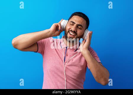 Junger, glücklicher arabischer Mann mit Kopfhörern, der vor blauem Hintergrund Musik hört Stockfoto