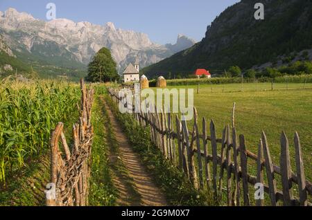 Ein Weg mit Holzzaun führt die katholische Kirche im Theth-Tal, im Hintergrund die Berge der albanischen Alpen Stockfoto