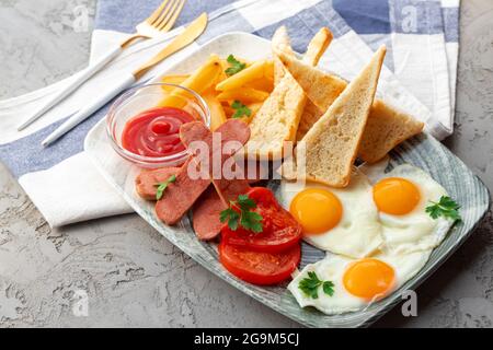 Englisches Frühstück mit Eiern, Würstchen, französischem Toast und Kartoffelkeilen auf einem grauen Tisch Stockfoto