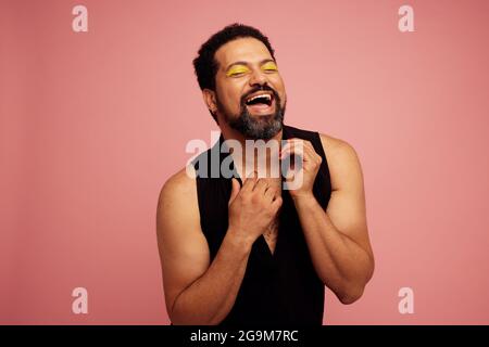 Grad Queen mit Augen Make-up lächelt auf rosa Hintergrund mit geschlossenen Augen. Genderqueer in schwarzem T-Shirt und Make-up sieht glücklich aus. Stockfoto