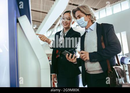 Eine weibliche Bodenbegleiterin mit Gesichtsschutz hilft männlichen Reisenden mit einem Check-in-Automaten am Flughafenterminal während einer Pandemie. Stockfoto