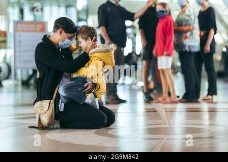 Frau und ihr Junge sitzen auf dem Boden am Flughafen. Mutter mit Gesichtsmaske, die mit ihrem Sohn am internationalen Flughafenterminal sitzt. Stockfoto