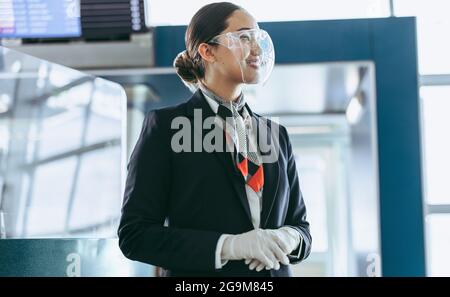 Lufthostess im Gesichtsschutz, der am Flughafen steht. Flughafenmitarbeiter am Terminal während einer Pandemie, die auf Passagiere wartet. Stockfoto