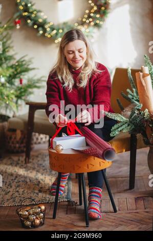 Lächelnd glücklich blonde Frau mittleren Alters sitzt auf Stuhl im neuen Jahr festlich dekoriert Wohnzimmer und Verpackung weihnachtsgeschenk-Box mit Geschenkpapier Stockfoto