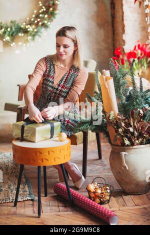 Lächelnde, fröhliche blonde Frau, die im neuen Jahr auf einem Stuhl sitzt, hat ein festlich dekoriertes Wohnzimmer und eine Geschenkschachtel mit Geschenkpapier und Band verpackt Stockfoto