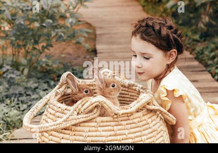 Kleines hübsches Mädchen im Sommerkleid, das draußen auf einem Holzweg vor dem Strohkorb mit Kaninchen sitzt, niedliches Kind, das mit einem leichten Lächeln Hasen ansieht Stockfoto