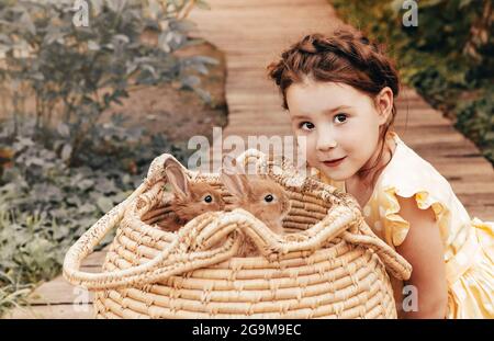 Kleines hübsches Mädchen im Sommerkleid, das draußen auf einem Holzweg vor dem Strohkorb mit Kaninchen sitzt, niedliches Kind, das mit einem leichten Lächeln Hasen ansieht Stockfoto