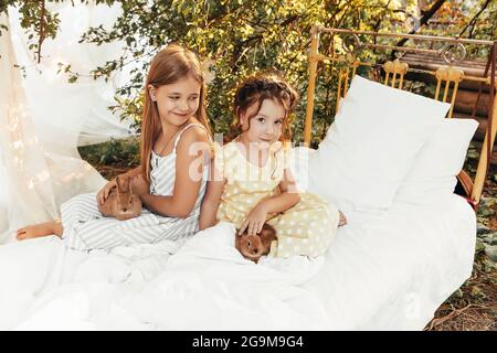 Kleine hübsche Mädchen in Sommerkleidern sitzen draußen zusammen mit Kaninchen, niedliche Kinder Blick auf Hasen mit leichtem Lächeln genießen die Zeit mit Stockfoto