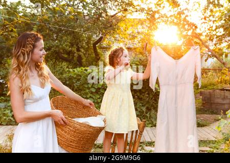 Charming kleines Mädchen helfen Mutter mit Weidenkorb während der Arbeit Chore und hängende Wäsche im Hinterhof im Sommer Abend Stockfoto