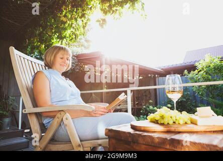 Eine fröhliche Frau mittleren Alters mit einem Glas Weißwein, die lächelte und das Buch las, während sie auf der Veranda an der Glaswand des Hauses saß Stockfoto