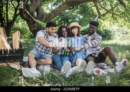 Junge, fröhliche Männer und Frauen sitzen im Sommergarten auf kariertem Karli zusammen und trinken kaltes Bier. Multikulturelle Freunde genießen Picknick-Zeit an der frischen Luft. Stockfoto