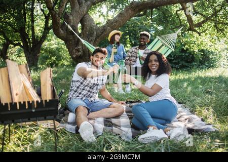 Glückliche junge, multiethnische Menschen chillen gemeinsam im grünen Sommerpark. Zwei Paare in legerer Kleidung sitzen auf Gras, entspannen sich in der Hängematte und trinken Bier. Stockfoto
