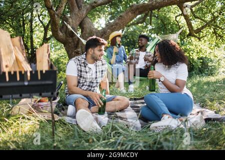 Eine Gruppe von vier fröhlichen Freunden in legerer Kleidung, die im Sommergarten picknicken. Zwei multirassische Paare sitzen auf Plaid und Hängematte, reden und trinken Alkohol. Stockfoto