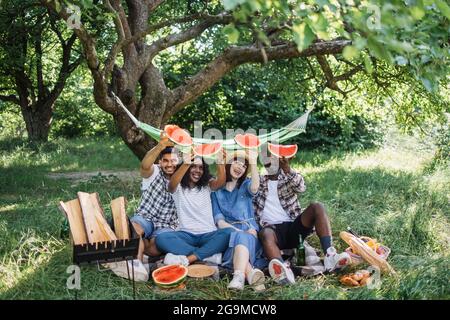 Fröhliche Männer und Frauen, die auf einer karierten Decke im Garten sitzen und Wassermelonenscheiben halten. Sommerpicknick von vier multirassischen Freunden. Stockfoto