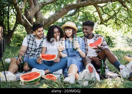 Gruppe von vier multirassischen Freunden, die ein modernes Smartphone benutzen, Wassermelone essen und Bier trinken. Glückliche junge Menschen, die ihre Freizeit in der Natur verbringen. Picknick im Garten im Sommer. Stockfoto