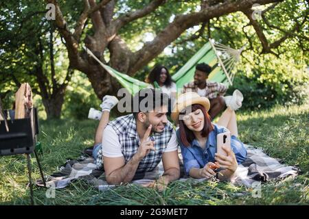 Fröhliches Paar beim Selfie auf dem Smartphone während des Picknicks im Freien. Verwischen Sie den Hintergrund des afrikanischen Mannes und der Frau, die in der Hängematte sitzen und Bier trinken. Summertime und Freundschaftskonzept. Stockfoto