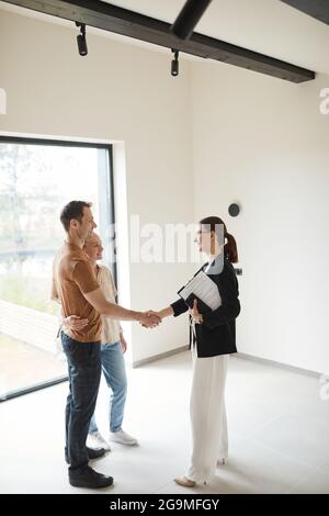 Vertikale Seitenansicht Porträt eines jungen Paares, das sich während der Tour durch die Wohnung mit dem Immobilienmakler die Hände schüttelt, Platz kopieren Stockfoto