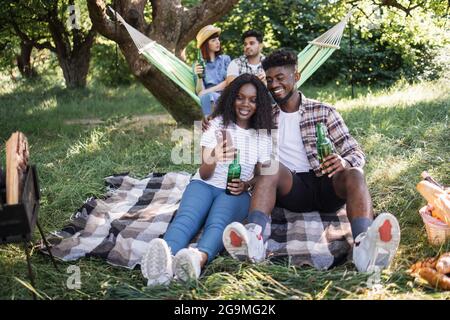 Schönes schwarzes Paar, das in Umarmungen im Garten sitzt und auf dem mobilen Bildschirm schaut, während ihre vielfältigen Freunde in der Hängematte reden und trinken. Vier Freunde beim Picknick in der Natur. Stockfoto