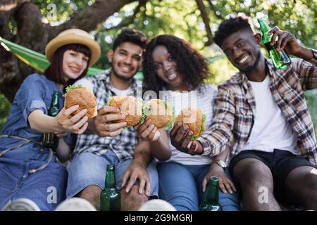 Positive multikulturelle Männer und Frauen sitzen auf weichem Plaid im grünen Garten und halten leckere frische Burger in den Händen. Junge glückliche Freunde beim Picknick mit Fast Food. Stockfoto