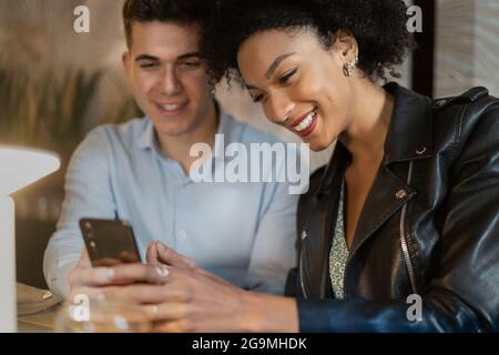 Lächelndes, multirassisches Paar junger Menschen, die ein Smartphone auf dem Tisch eines Restaurants benutzen. Menschen, die ein technisches Konzept nutzen. Stockfoto