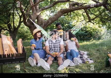 Drei junge Leute, die im Sommergarten auf Plaid sitzen und leckere Burger essen und Bier trinken. Verwischen Sie den Hintergrund eines afrikanischen Mannes, der in der Hängematte Gitarre spielt. Picknick von diversen Freunden. Stockfoto