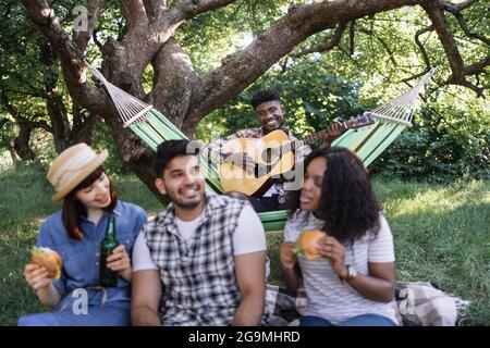 afroamerikanischer Kerl, der in der Hängematte sitzt und Gitarre spielt, während seine drei multikulturellen Freunde sich auf dem Gras mit Getränken und Snacks entspannen. Konzept von Mensch, Freizeit und Natur. Stockfoto